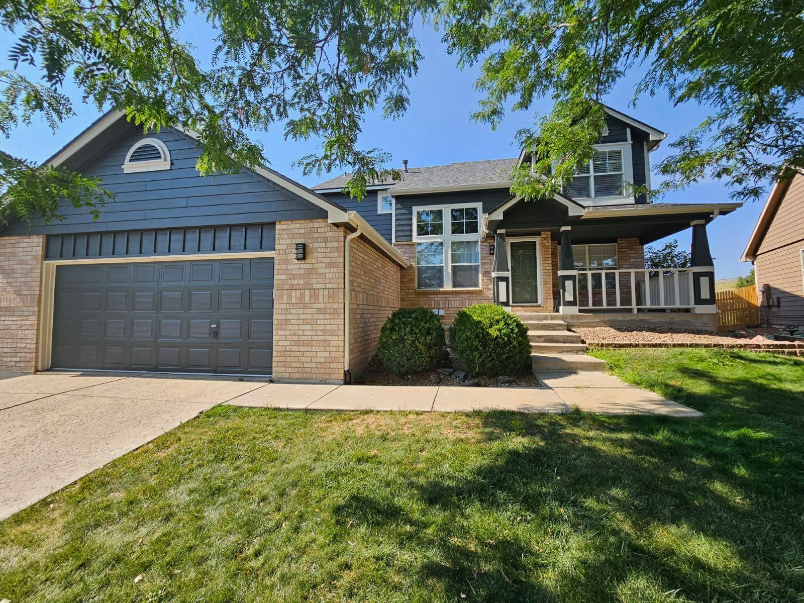Two-story house with dark blue siding, a brick facade, a two-car garage, and a front porch under a sunny blue sky.