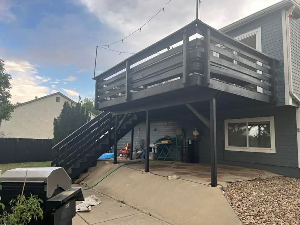 A dark-painted wooden deck with a staircase attached to the back of a grey house, overlooking a concrete patio and yard.