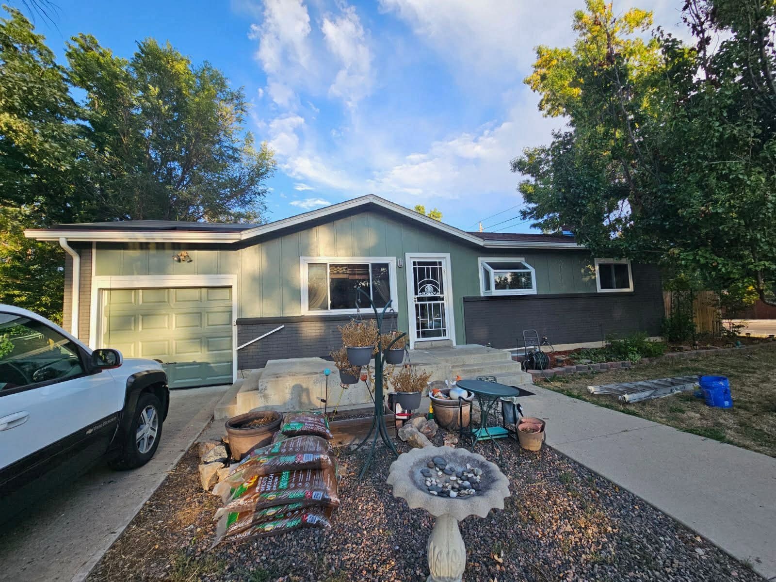 A light green house with a dark stone base, a white garage door, and a front yard with gravel, plants, and a birdbath.