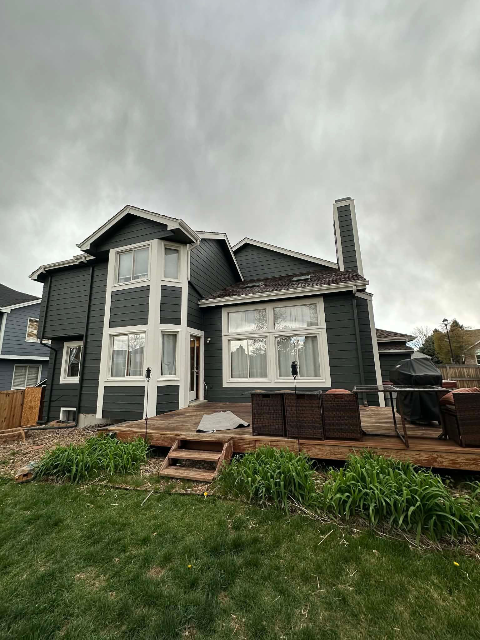 A two-story dark gray house with white window trim and a wooden deck under a cloudy sky.