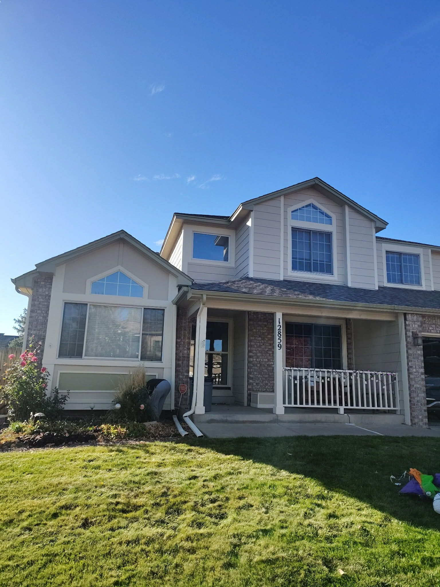 Two-story suburban home with tan siding, brick accents, and a front porch under a clear blue sky.
