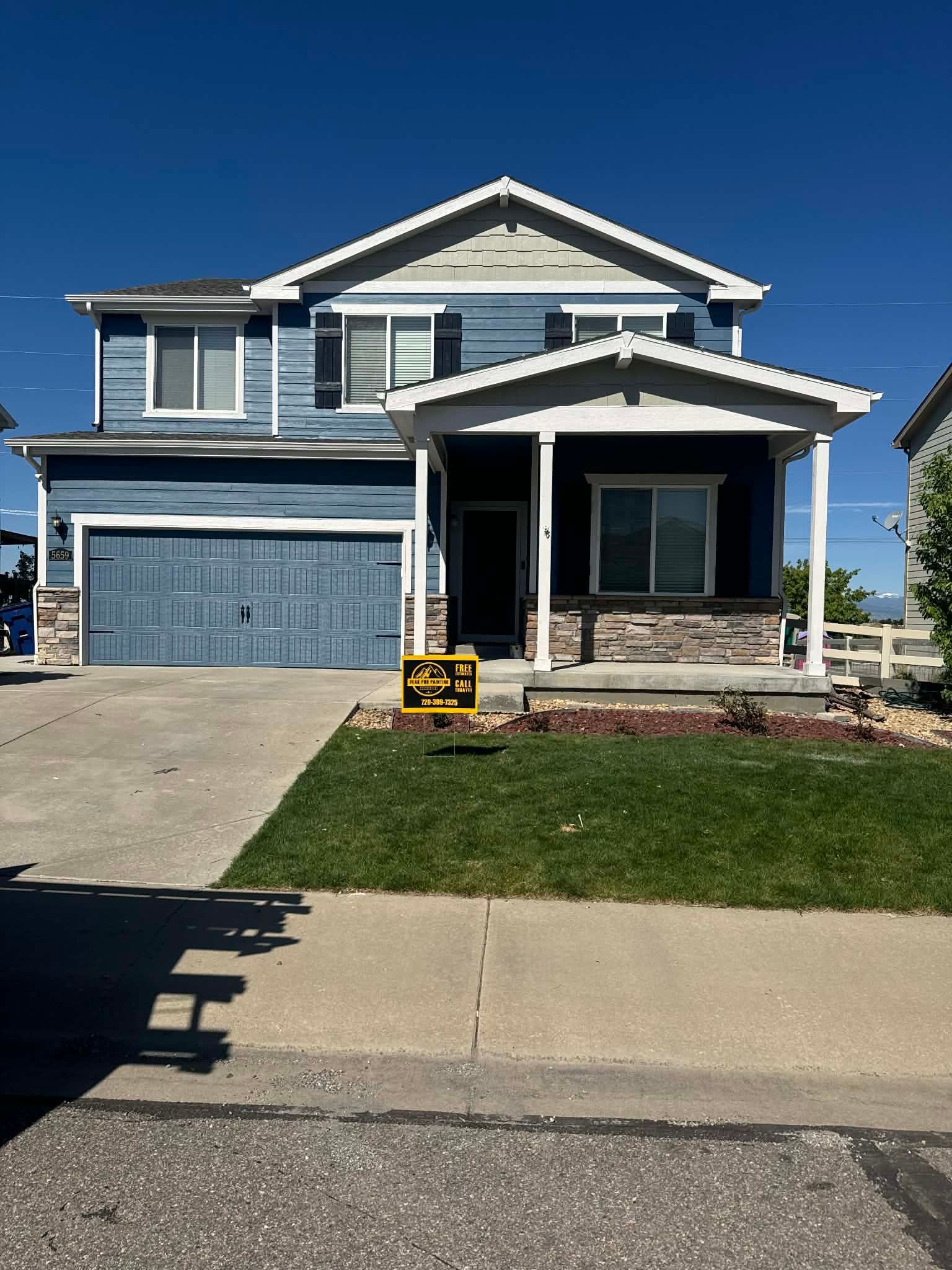 A two-story blue suburban house with a gray garage, front porch, and stone accents under a clear blue sky.