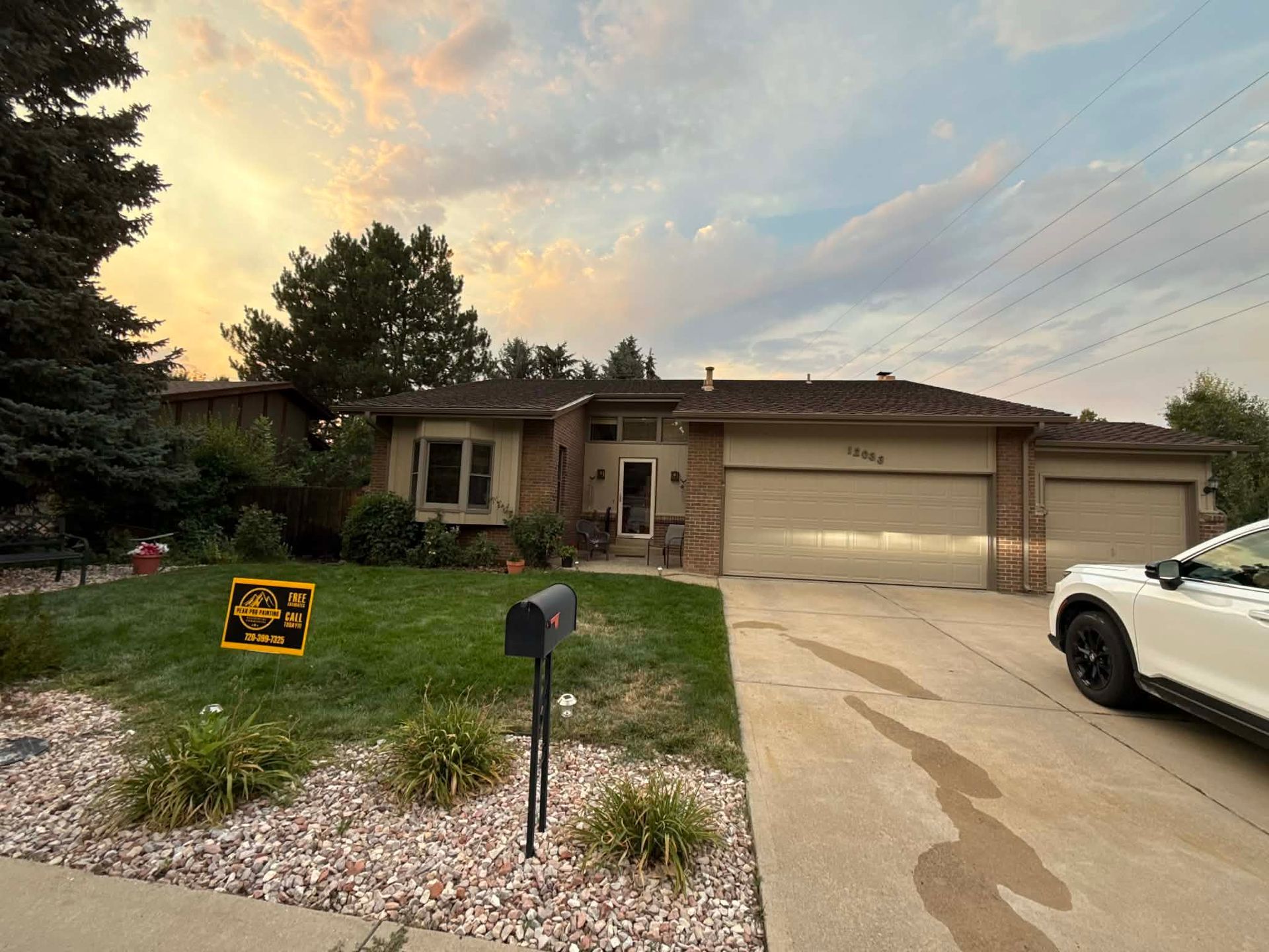 A single-story tan house with a brick entryway, attached garages, and a manicured front lawn at sunset.