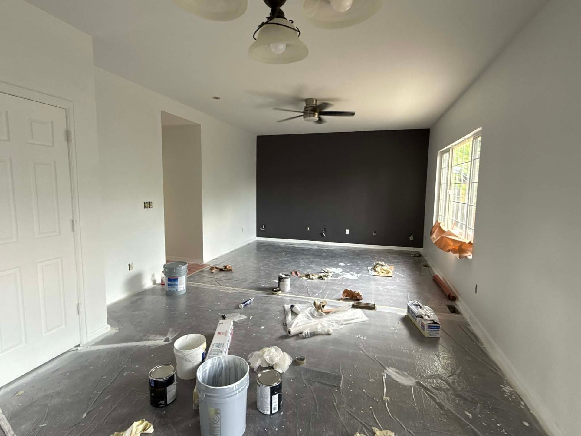 A room under renovation with white walls, a dark accent wall, a ceiling fan, and painting supplies spread on floor plastic.