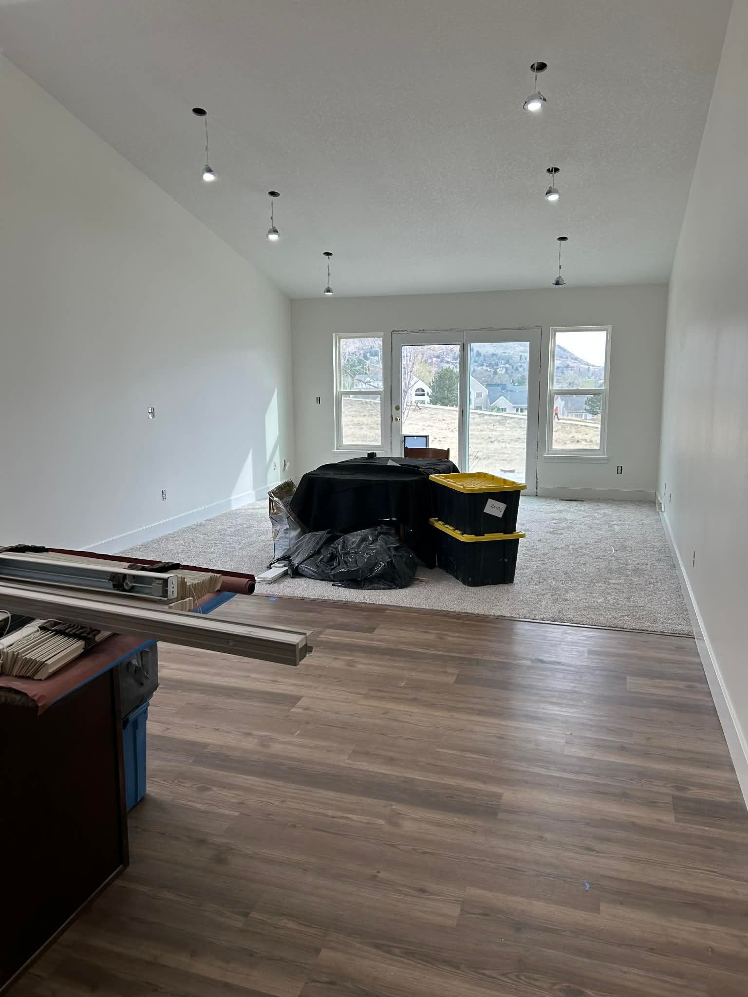An unfinished living room with wood-look floors, a patch of carpet, black storage bins, and bright overhead lighting.