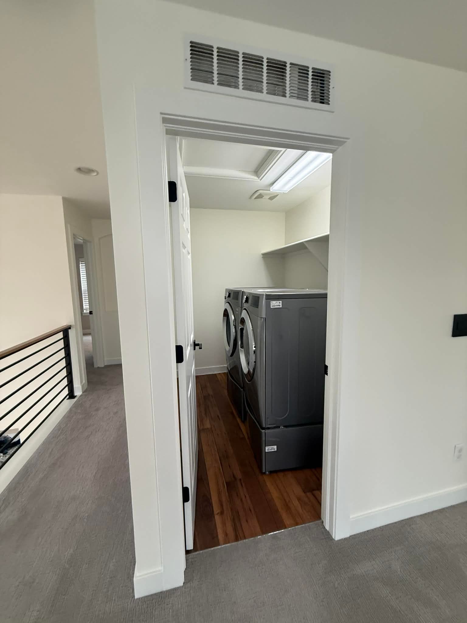A laundry closet containing two dark-gray machines, featuring a wooden floor and a white door frame in a carpeted hallway.