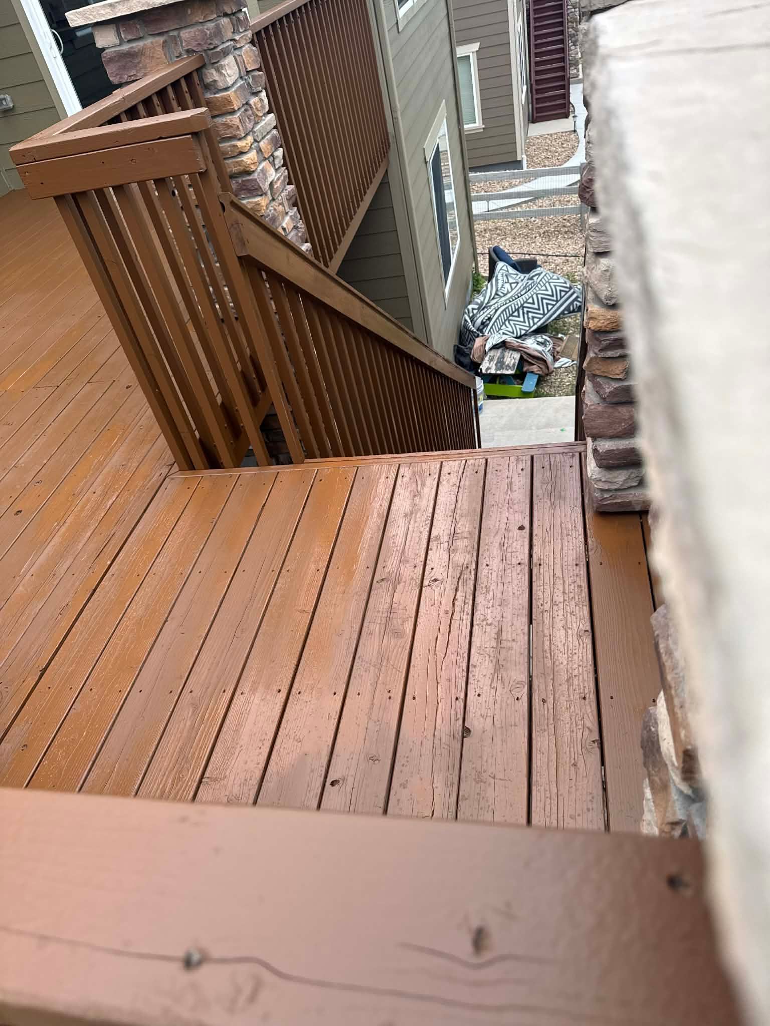 A brown wooden deck with stairs leading down to a side yard with a small, cluttered area against the house wall.