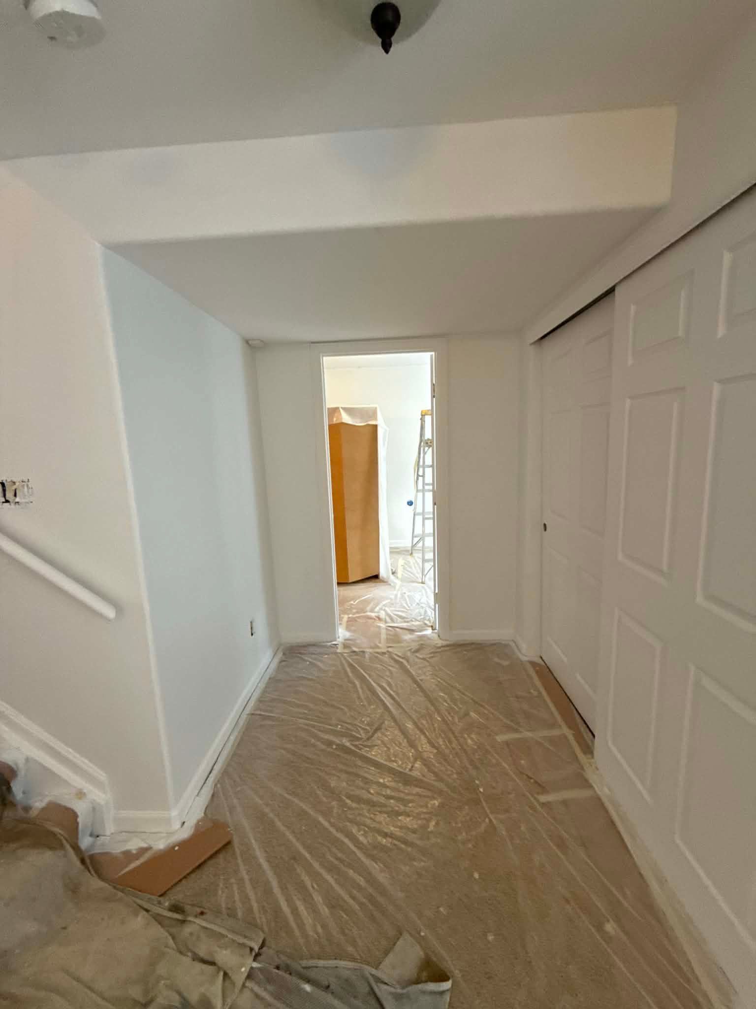 A hallway undergoing renovation, with white walls, plastic floor protection, and an open doorway to another room.