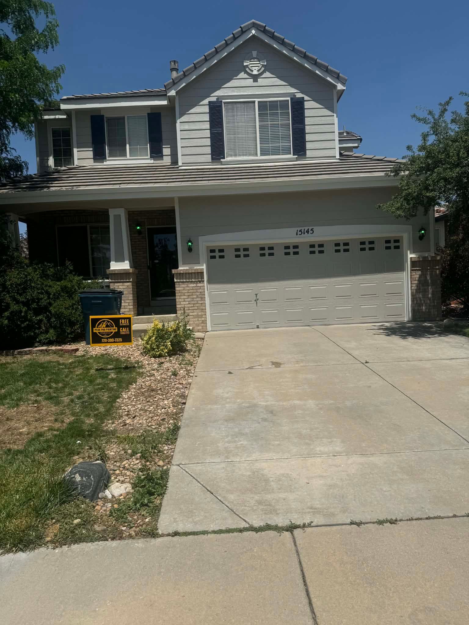 A two-story light gray house with a beige garage, dark shutters, stone accents, and a concrete driveway on a sunny day.