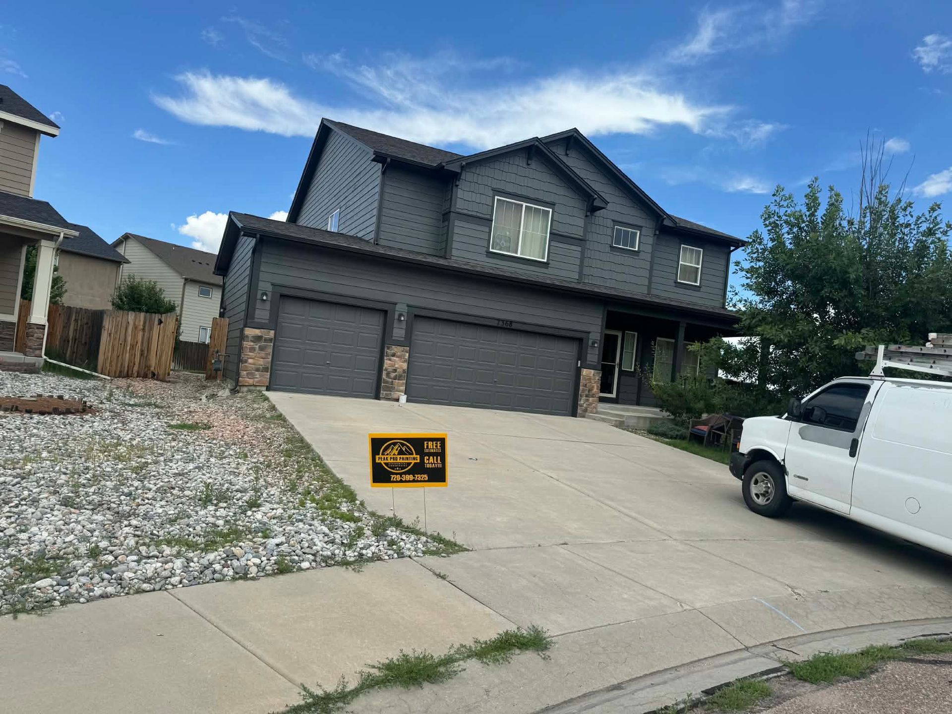 A two-story, dark-sided house with a double garage and a white van parked on the concrete driveway under a blue sky.
