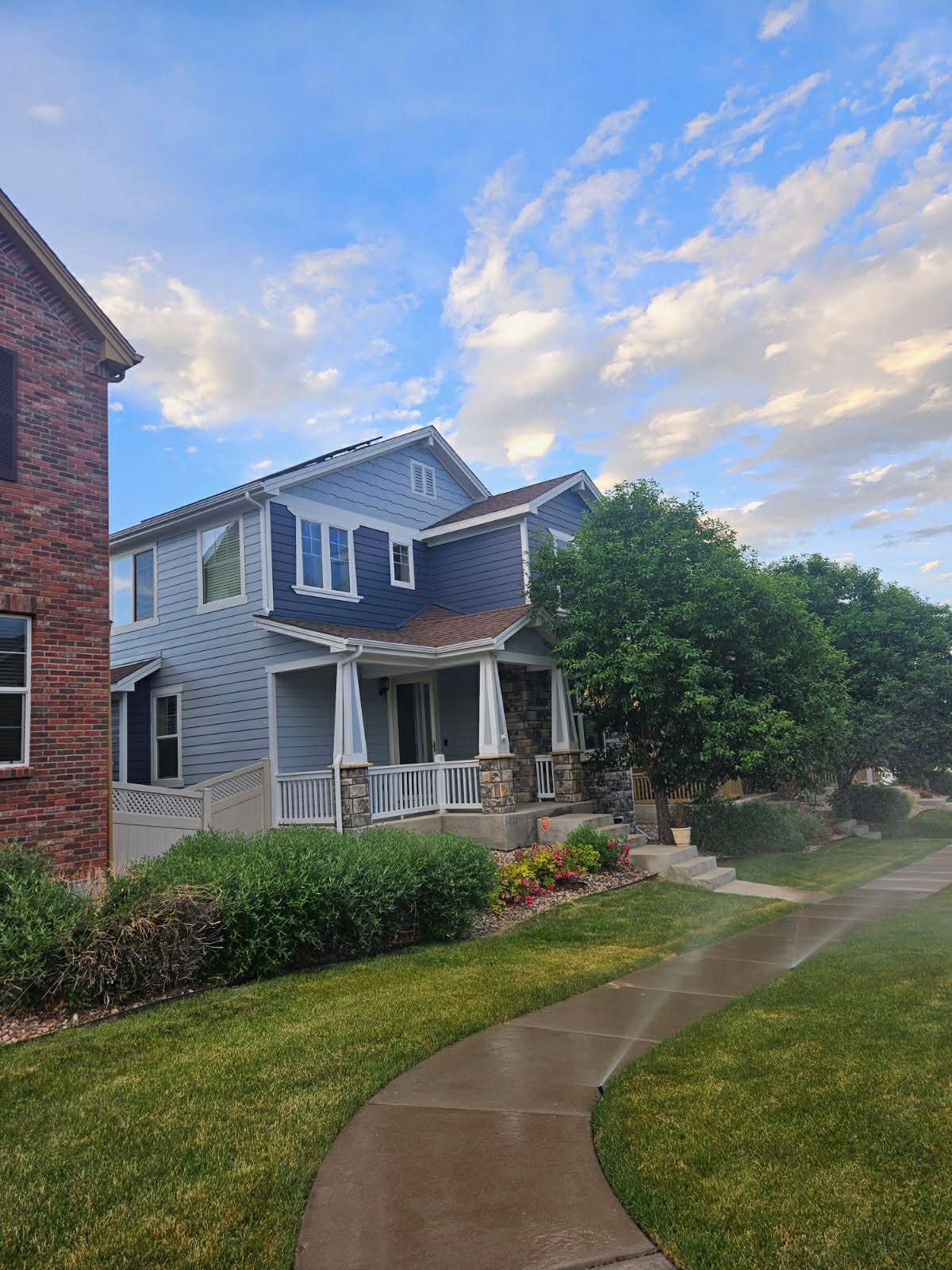 A blue two-story house with a front porch, stone accents, and manicured landscaping sits by a sidewalk under a cloudy sky.