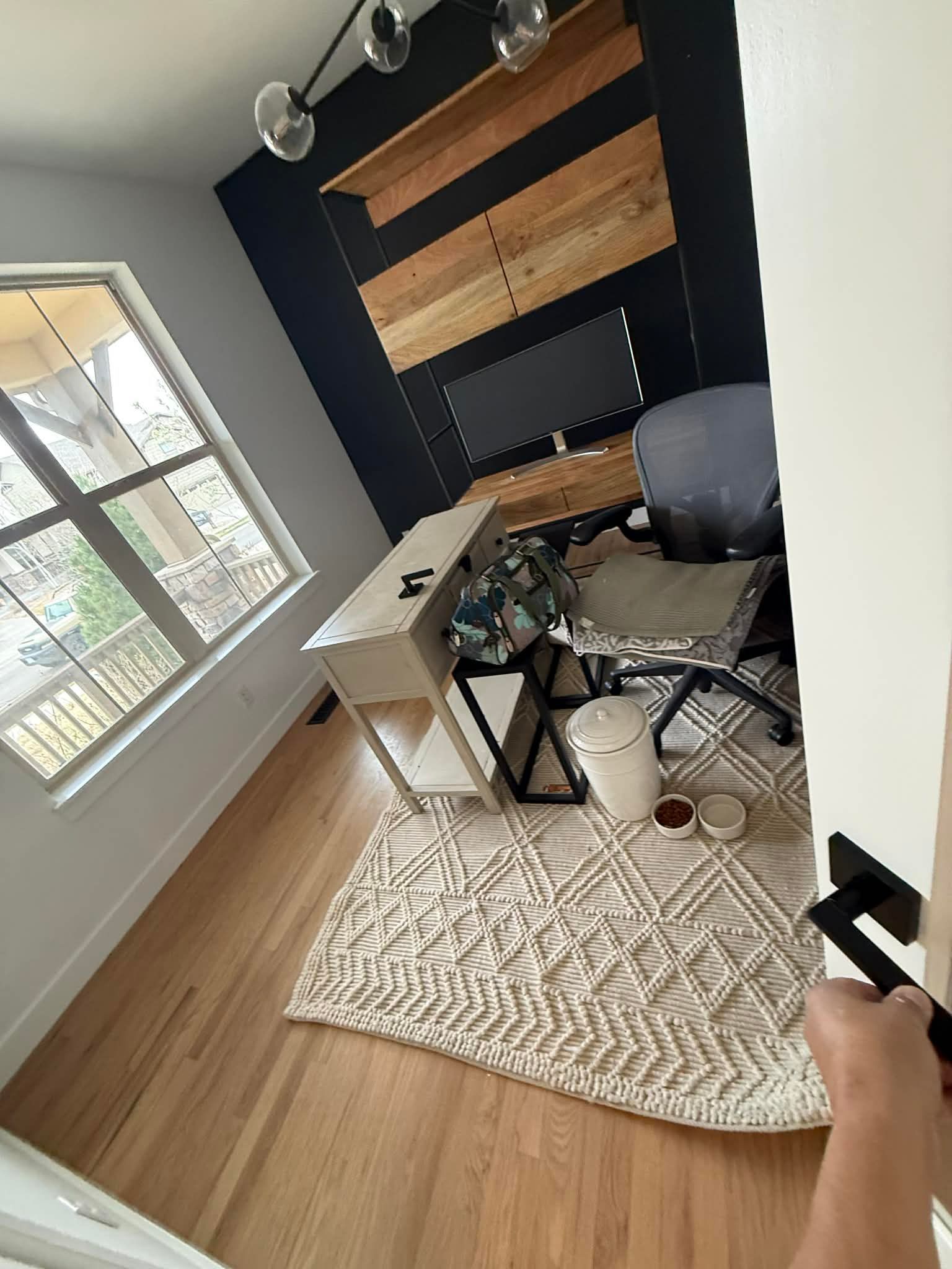 A home office featuring a desk with a monitor, a grey office chair, and wooden wall shelving on a textured rug.
