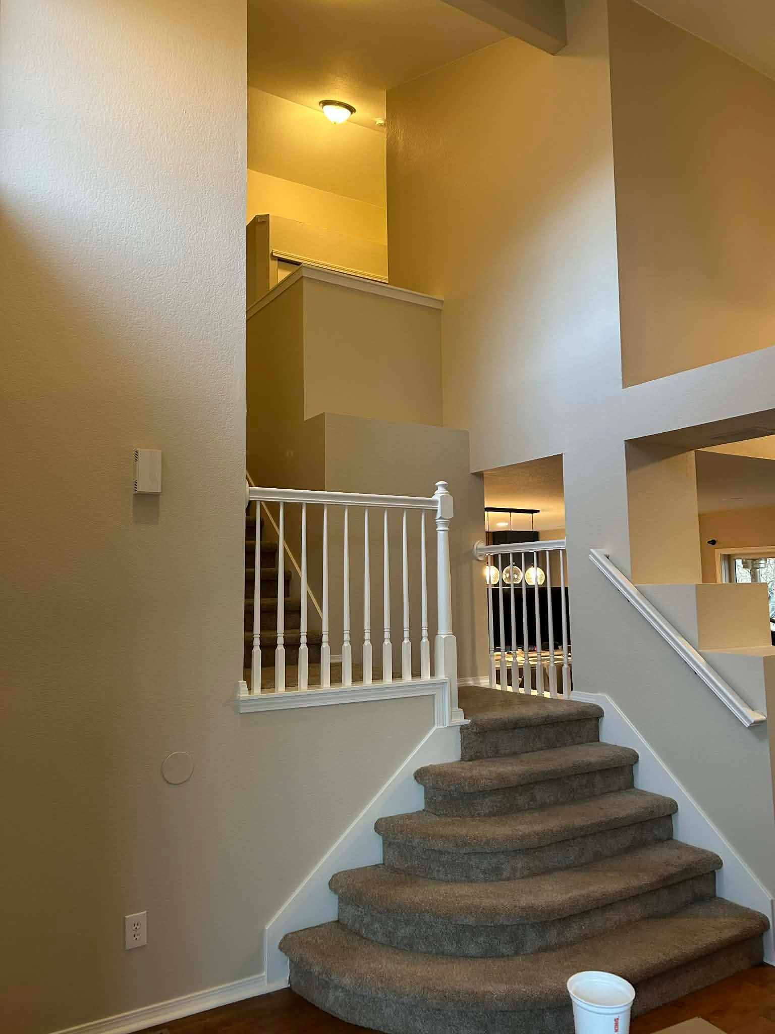 Carpeted stairs with white railings lead to an upper landing in a house with light beige walls and high ceilings.