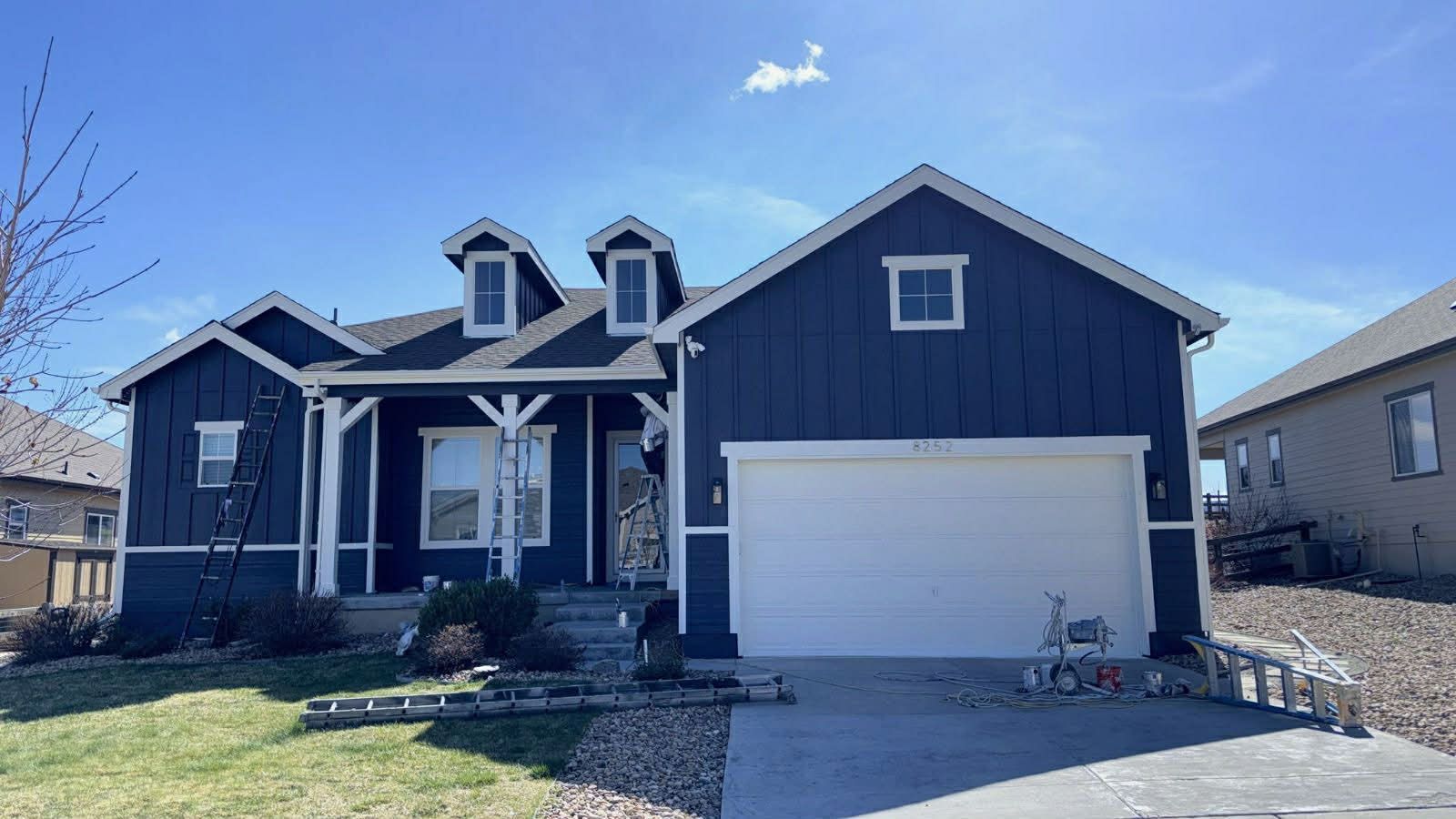 A one-story dark blue suburban house with a two-car garage, white trim, and two roof dormers under a bright blue sky.