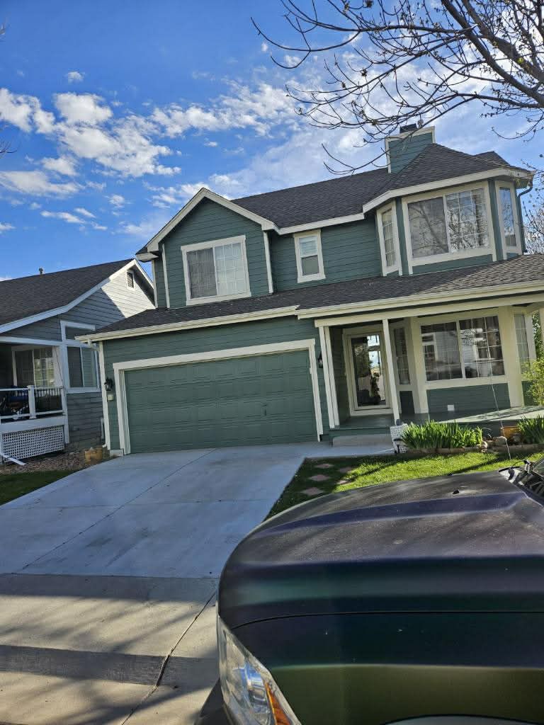 A two-story, sage green residential house with a large garage, concrete driveway, and a partially obscured vehicle.