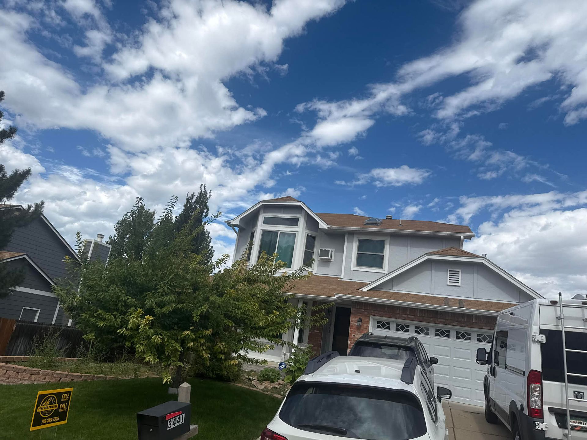 A two-story light gray house with a brown roof under a bright, cloudy blue sky, featuring a garage and driveway vehicles.