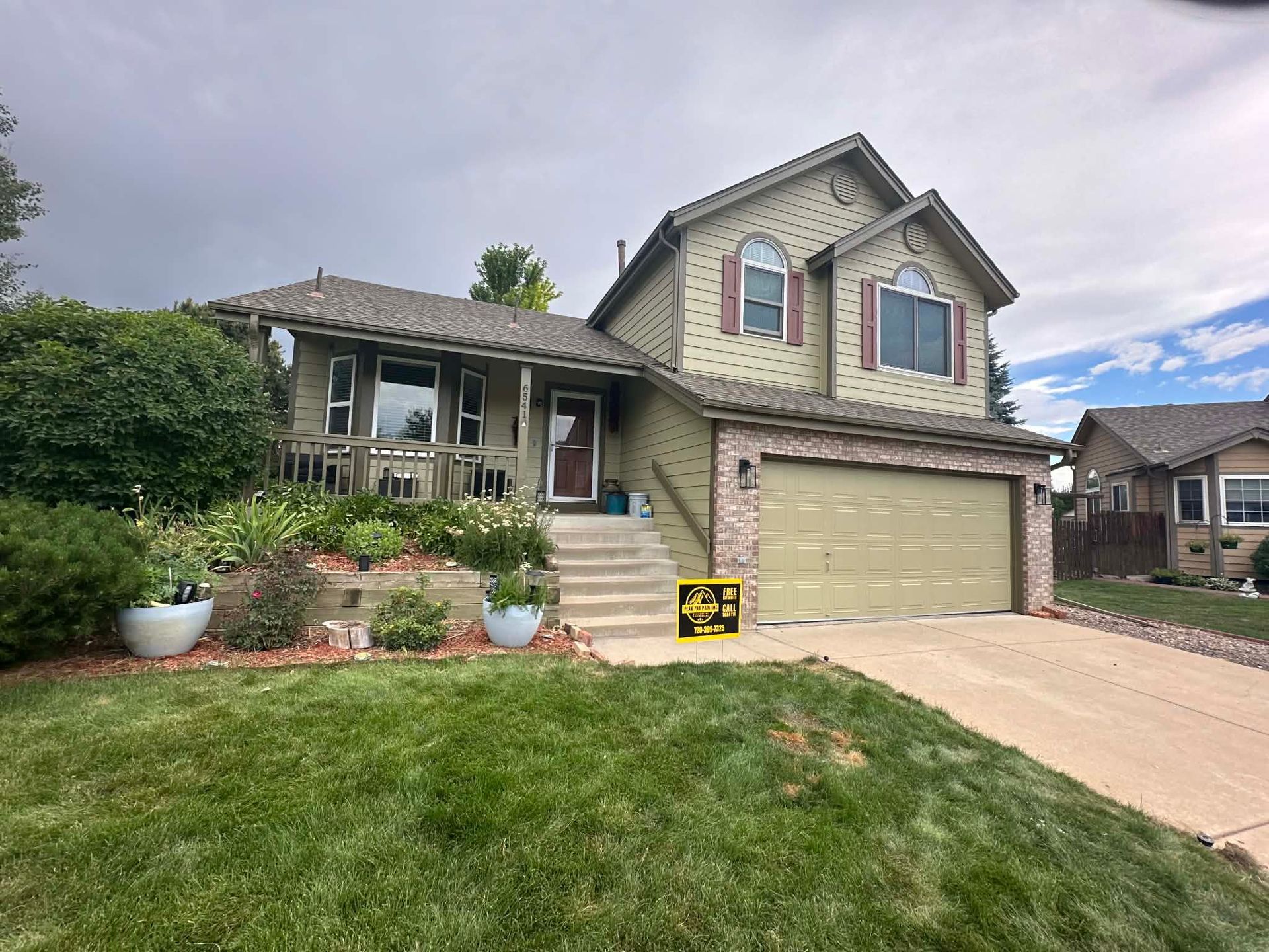 A two-story, tan-sided house with a brick garage, a concrete driveway, and a front yard with a landscaped garden.
