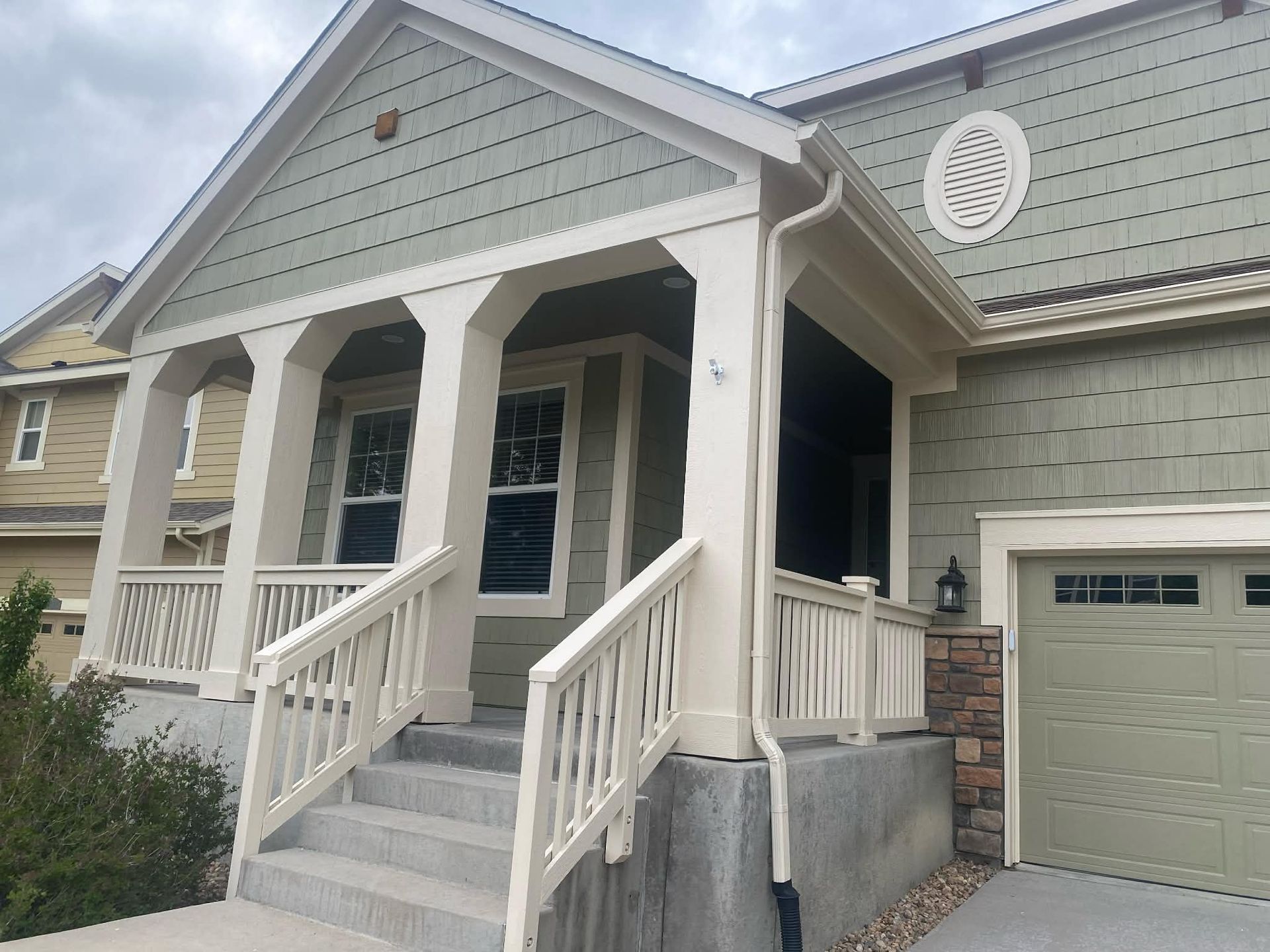 A light green suburban home with a covered front porch, white railings, arched support pillars, and a tan garage door.