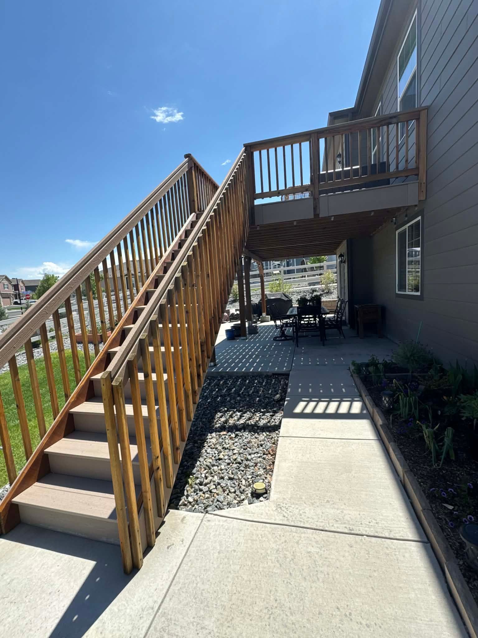 Wooden outdoor staircase leading to an elevated deck next to a gray house, with a concrete walkway below on a sunny day.