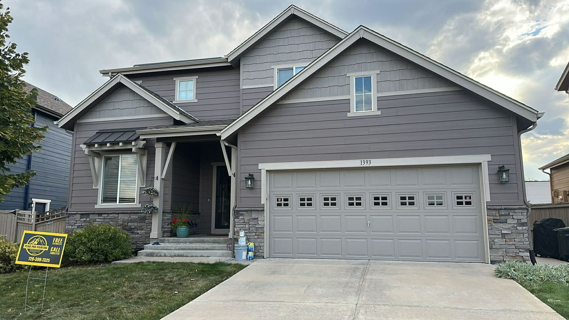A gray two-story suburban house with a two-car garage, stone masonry base, and a small front lawn under a cloudy sky.