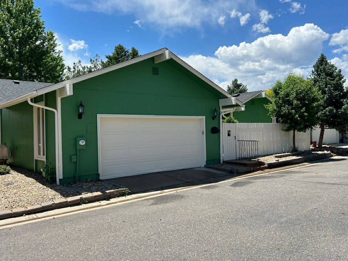 A bright green house with a white garage door, white trim, and a white fence under a sunny, blue sky with clouds.