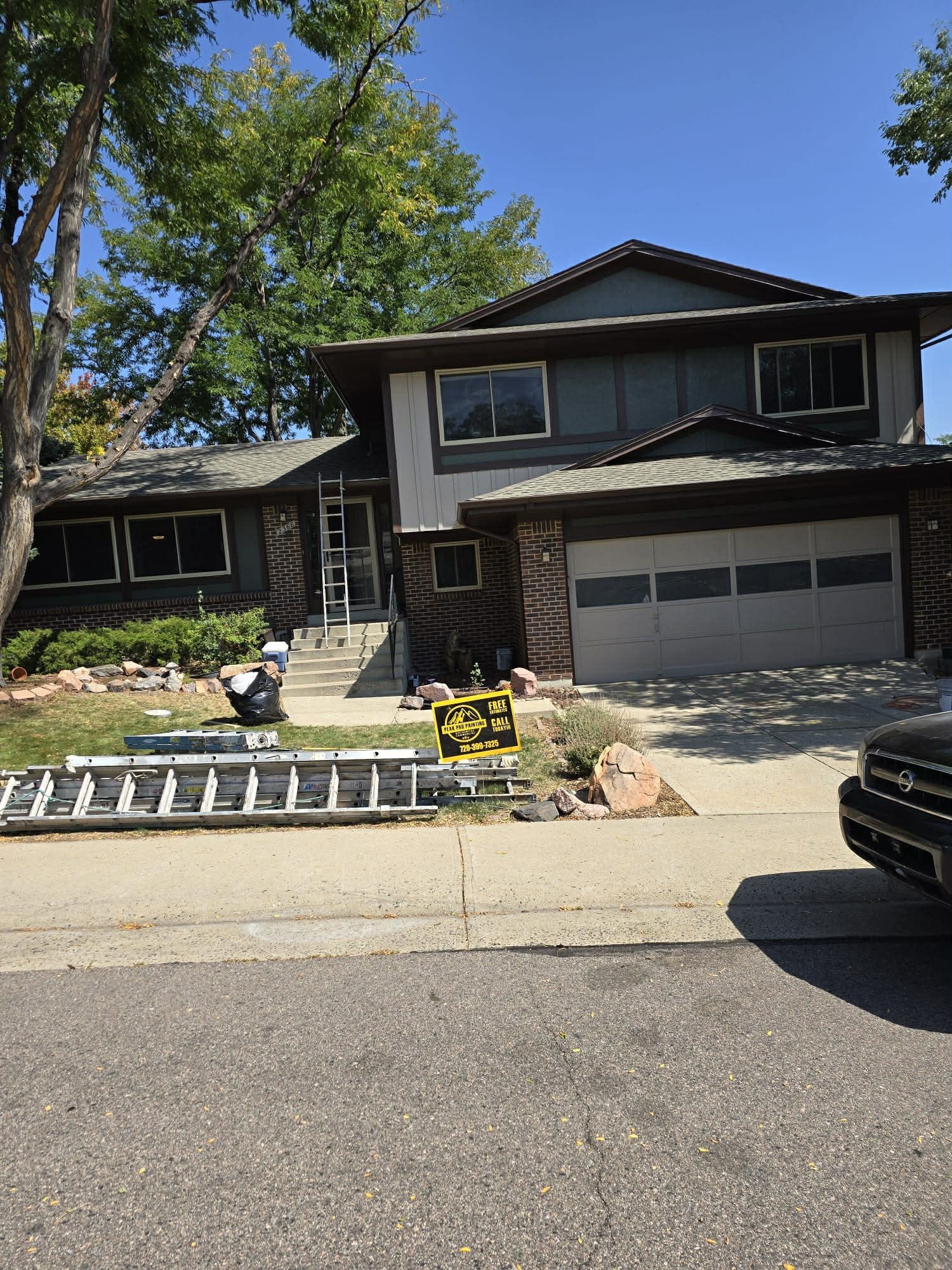 A two-story house with dark gray siding, brick trim, a two-car garage, and construction ladders on the front lawn.