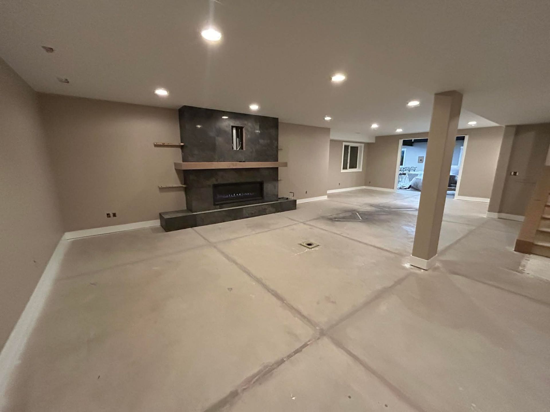 A spacious, unfinished basement with a modern dark stone fireplace, tan walls, recessed lighting, and a concrete floor.