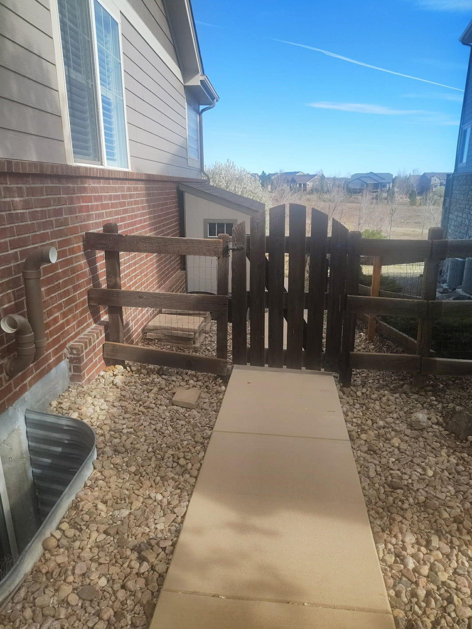 A concrete path leads to a wooden gate between a brick house and a fence, with gravel ground under a clear blue sky.