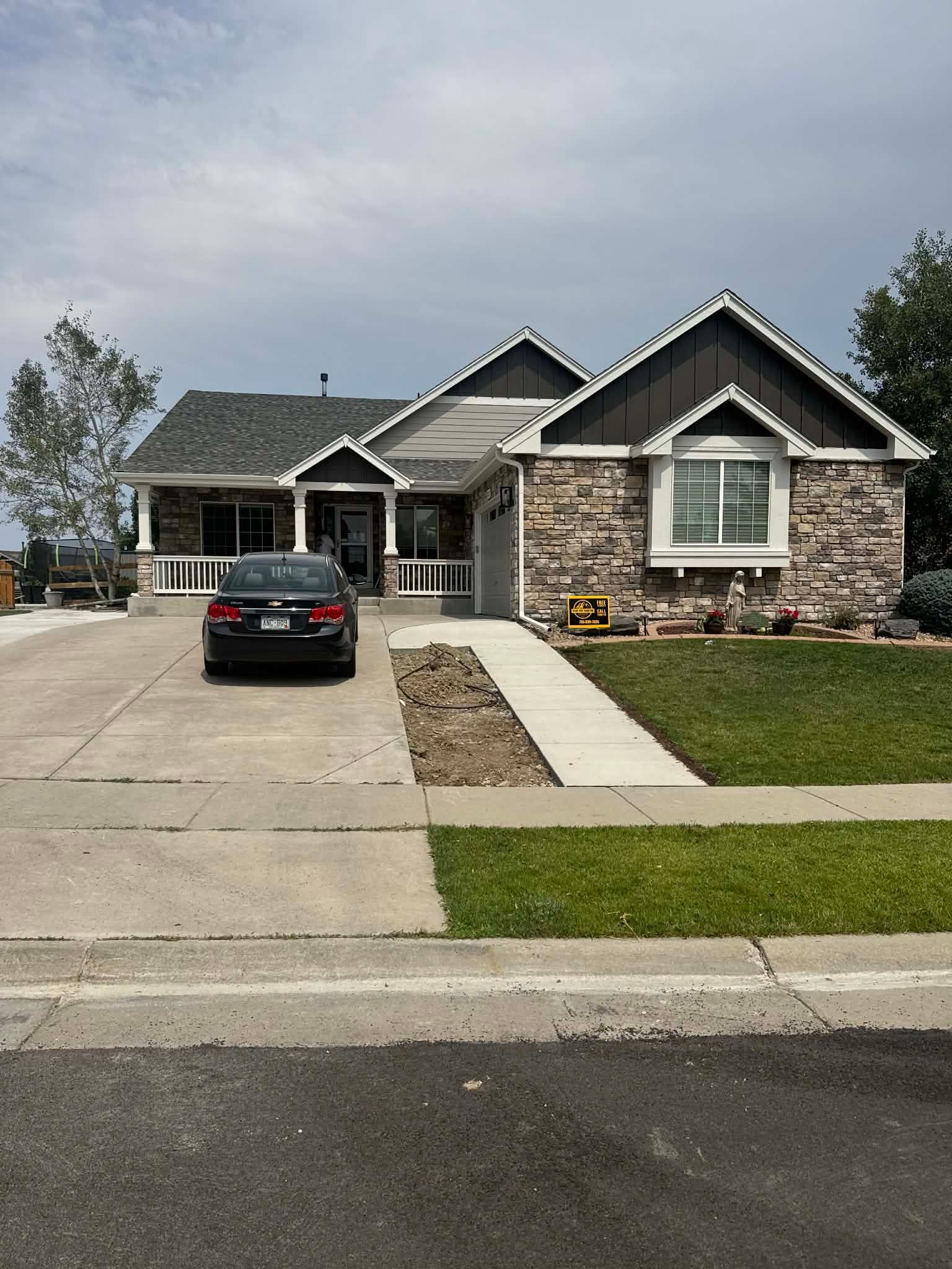 A single-story house with stone siding, a dark roof, and a driveway with a car parked on it under a cloudy sky.