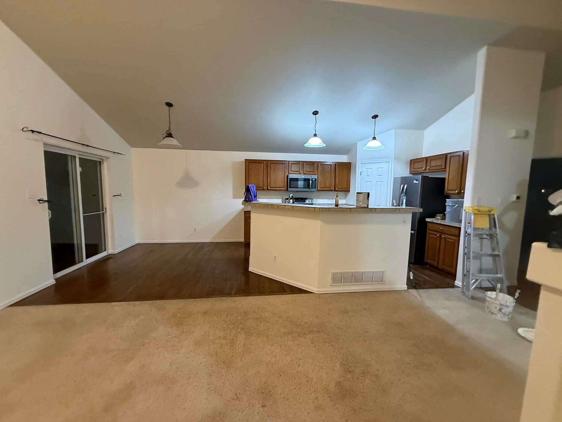 An open-concept living area with tan carpet transitioning to dark wood floors, a central kitchen island, and white walls.