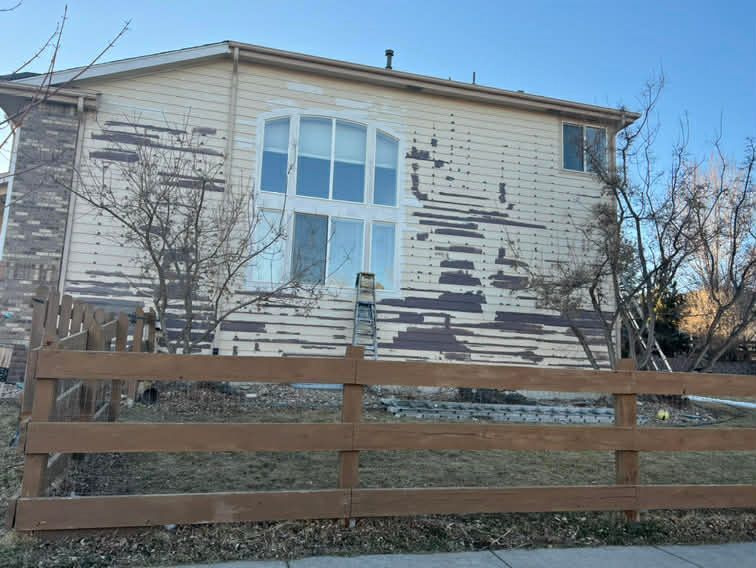 A two-story house with tan siding that is severely peeling, seen behind a wooden fence on a sunny day.