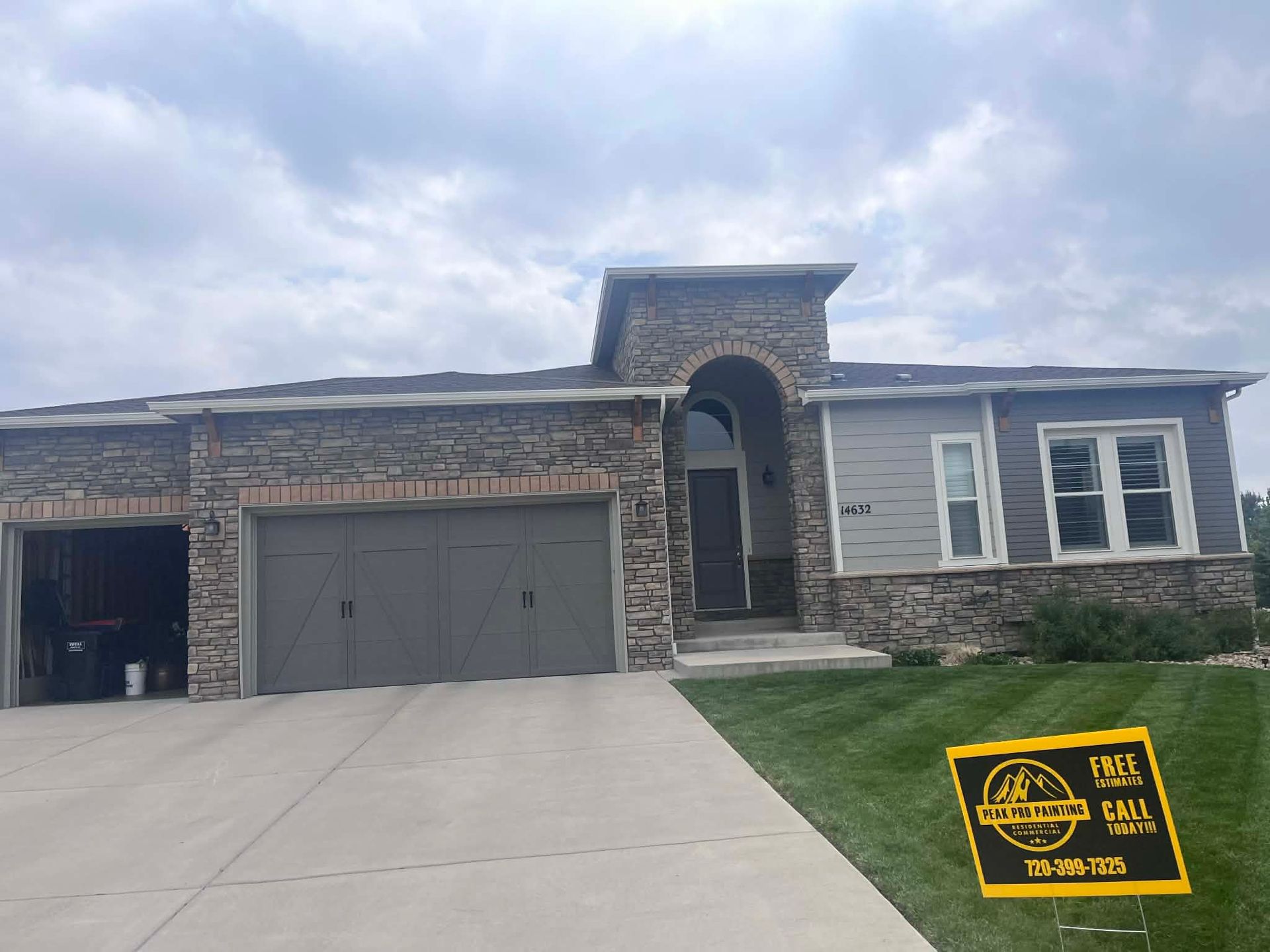 A single-story suburban house with stone and gray siding, a two-car garage, and a yard sign on a bright day.