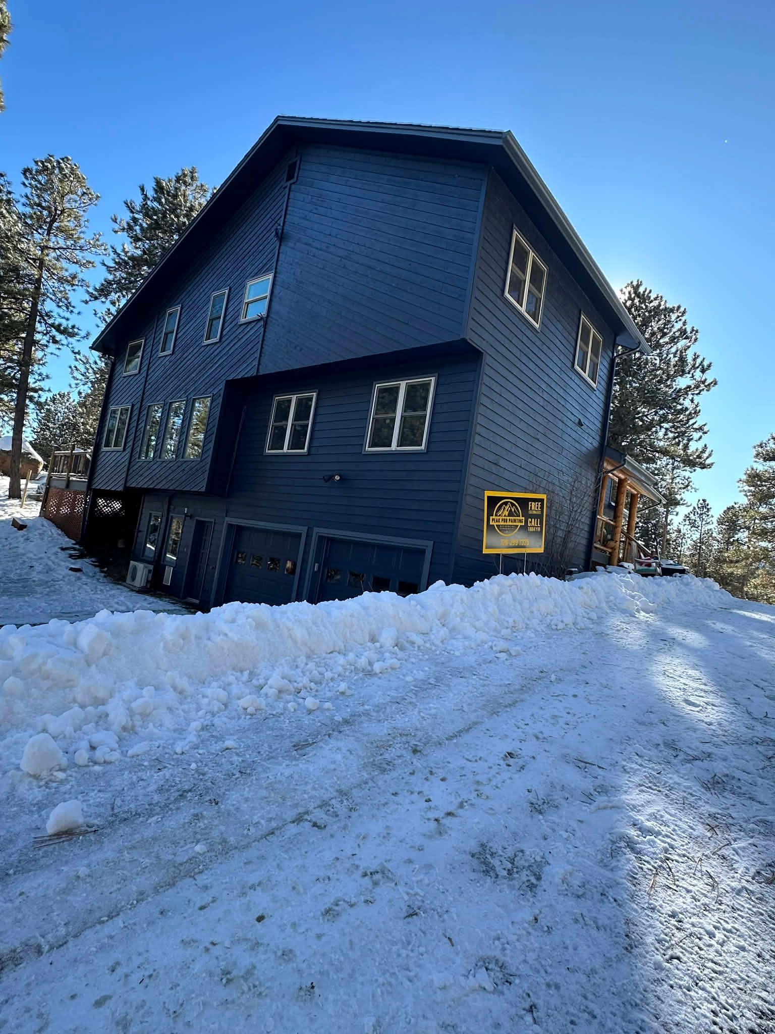 A dark-sided, multi-story house with a garage sits on a snowy, sloped lot under a clear blue sky.