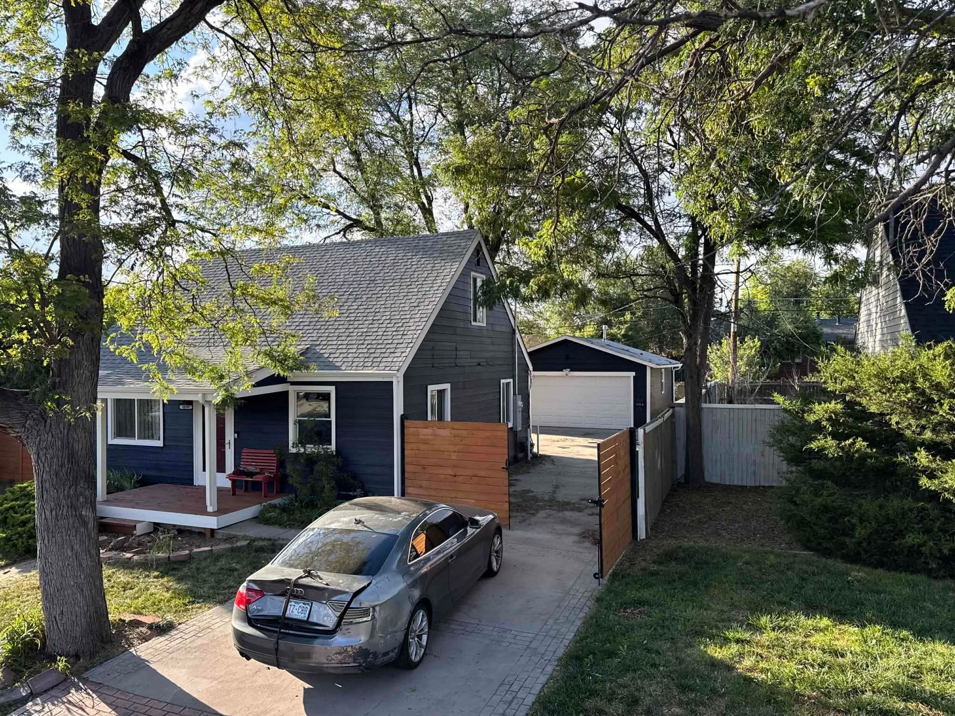 A dark-sided house with a front porch, a wood fence, and a gray car parked in the driveway leading to a detached garage.