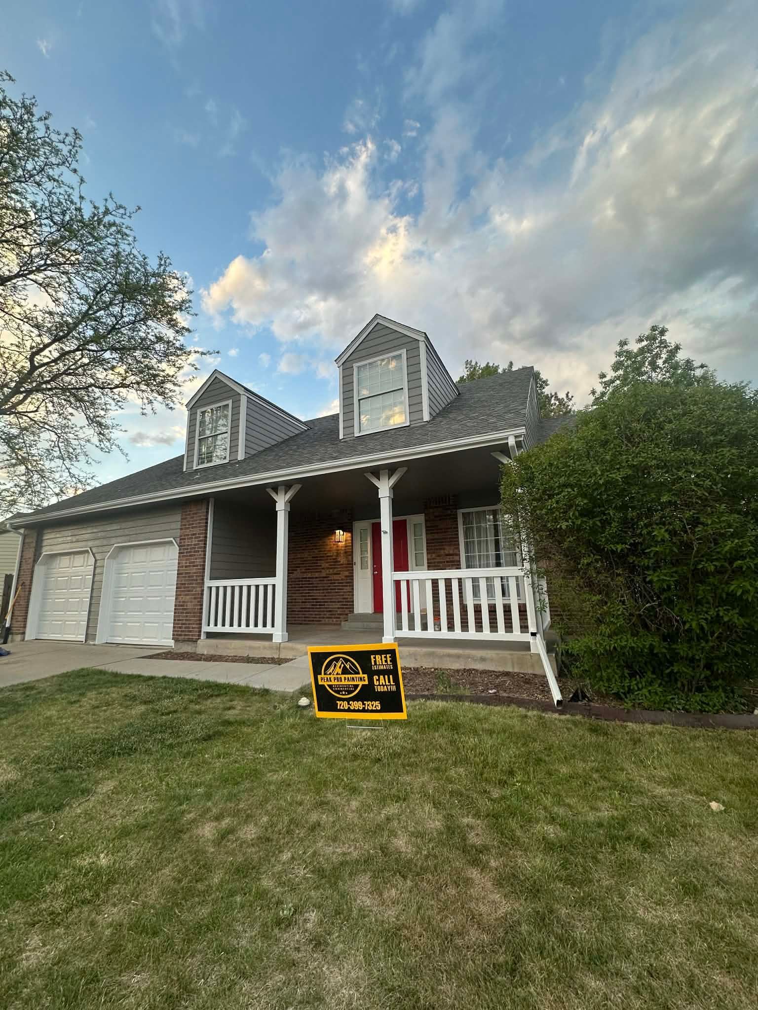 A house with a brick exterior, white porch railing, and a sign in the front yard under a blue sky with soft clouds.