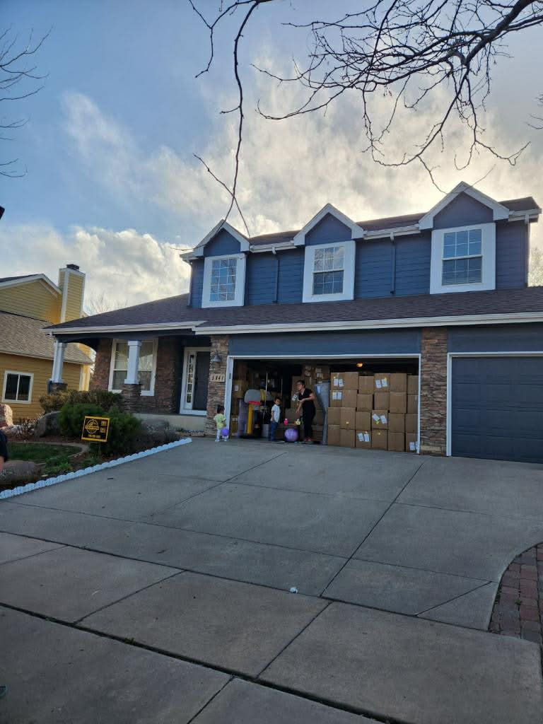 A blue two-story house with a driveway full of cardboard boxes and people standing in the open garage.