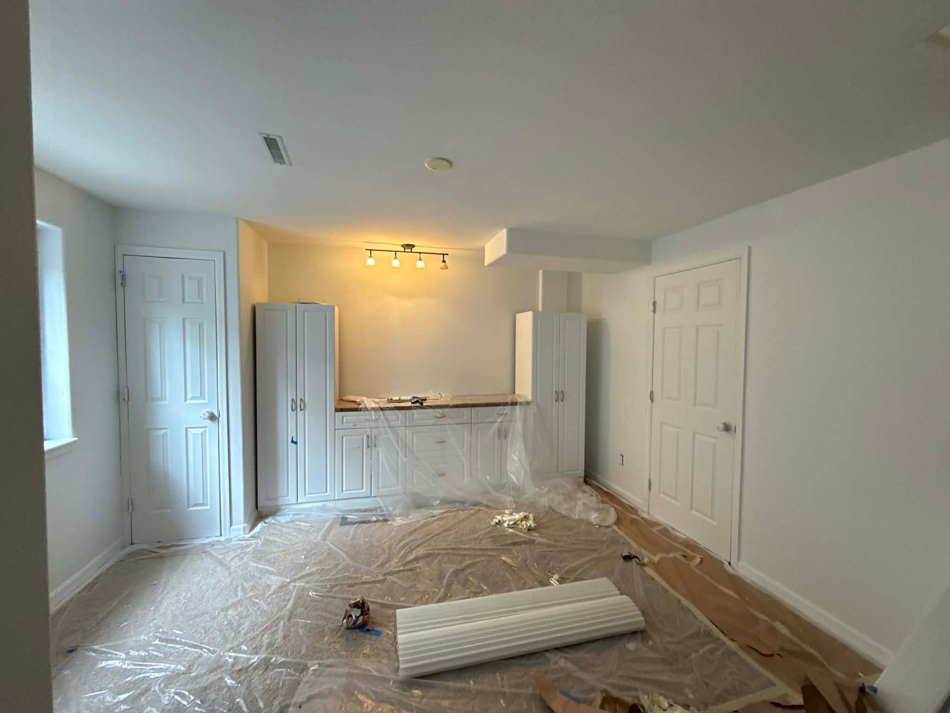 A white room under renovation with kitchen cabinetry installed, plastic floor coverings, and a central light fixture.