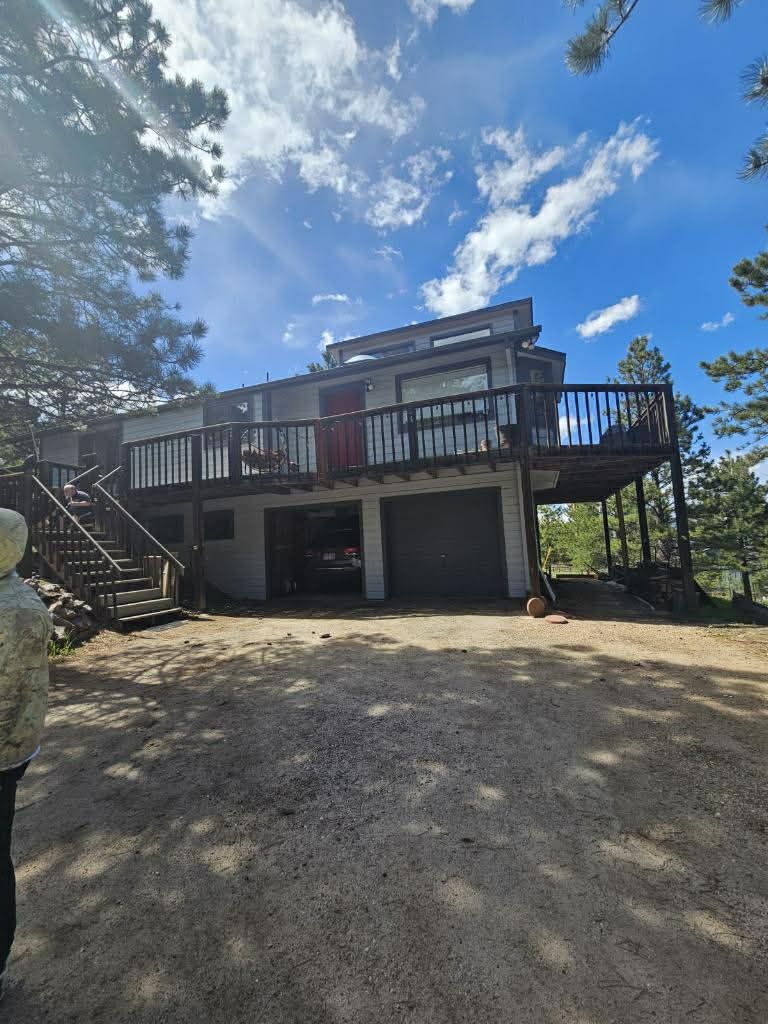 A two-story house with a wooden deck and attached garage stands on a gravel driveway under a bright blue sky.