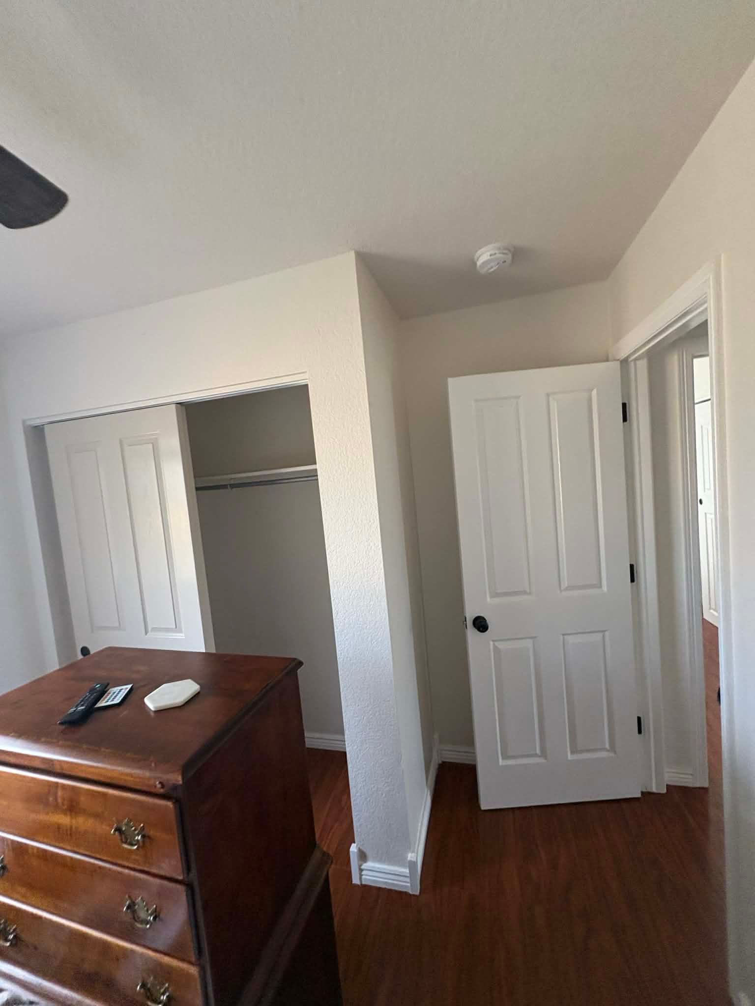 A bedroom corner featuring a wooden dresser in the foreground, an open closet, and a closed white door.