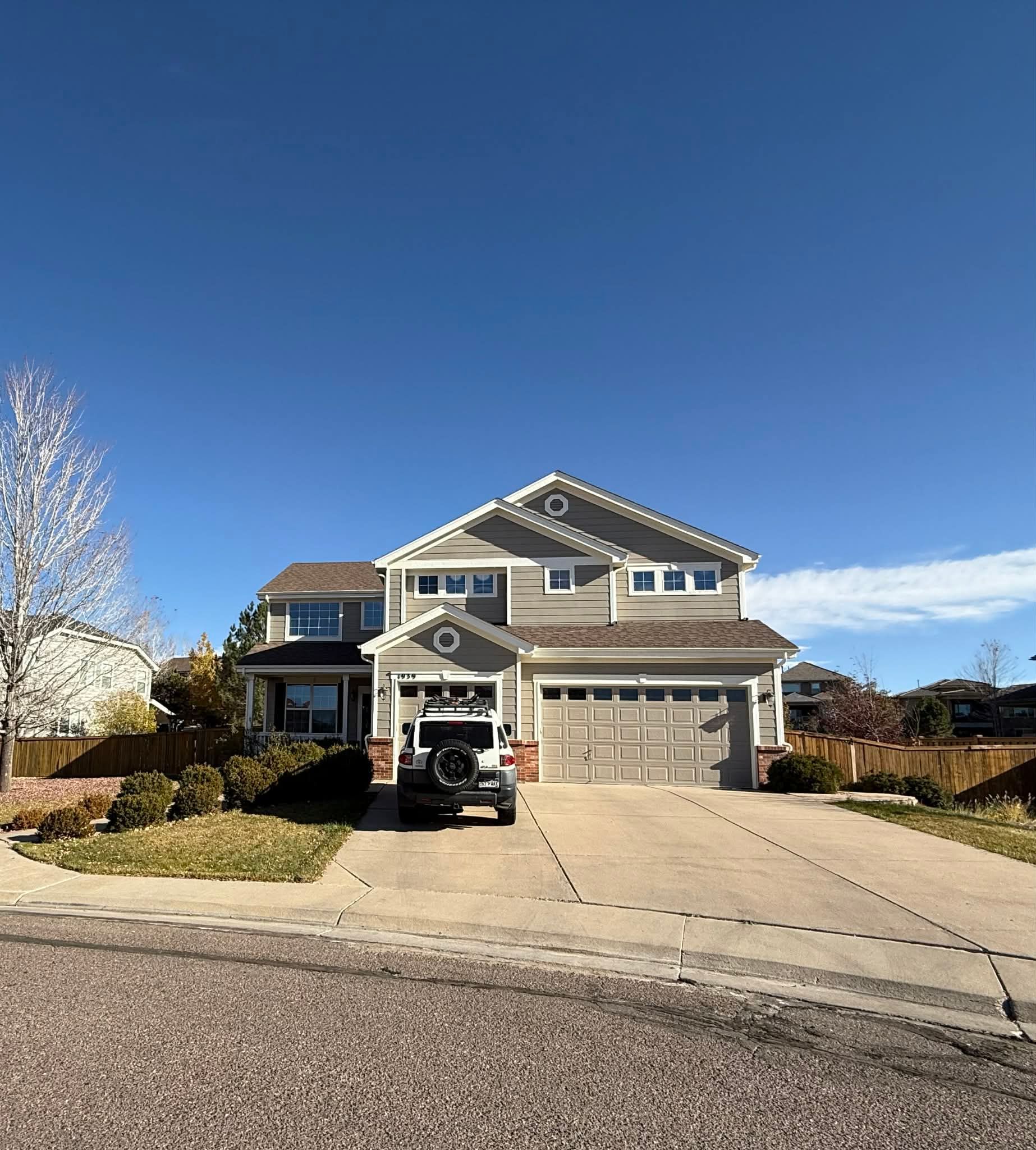 A two-story tan suburban house with a gray garage and a parked SUV in the driveway under a clear blue sky.