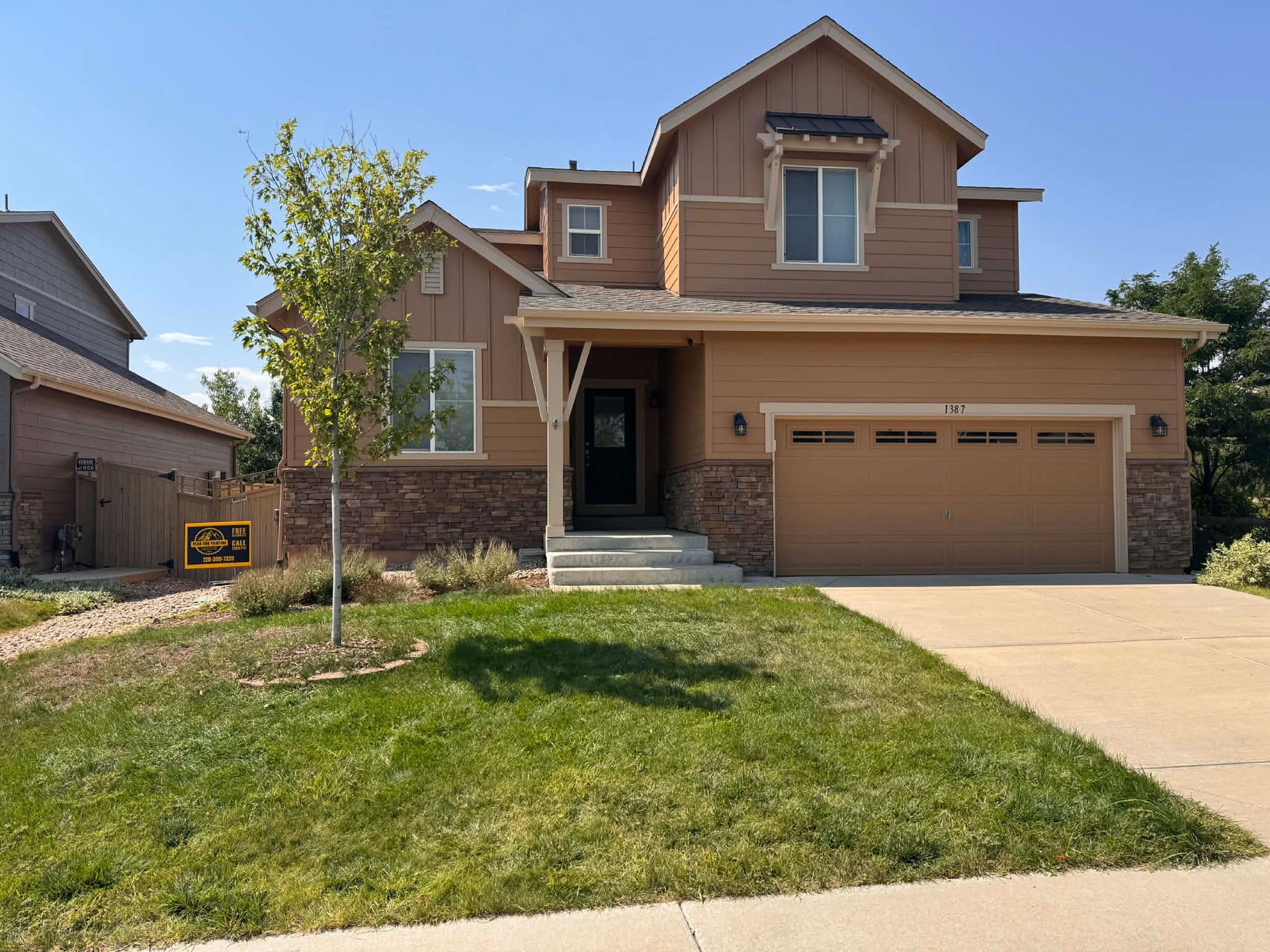 A two-story suburban home with light brown siding, stone accents, a two-car garage, and a small front yard with a tree.