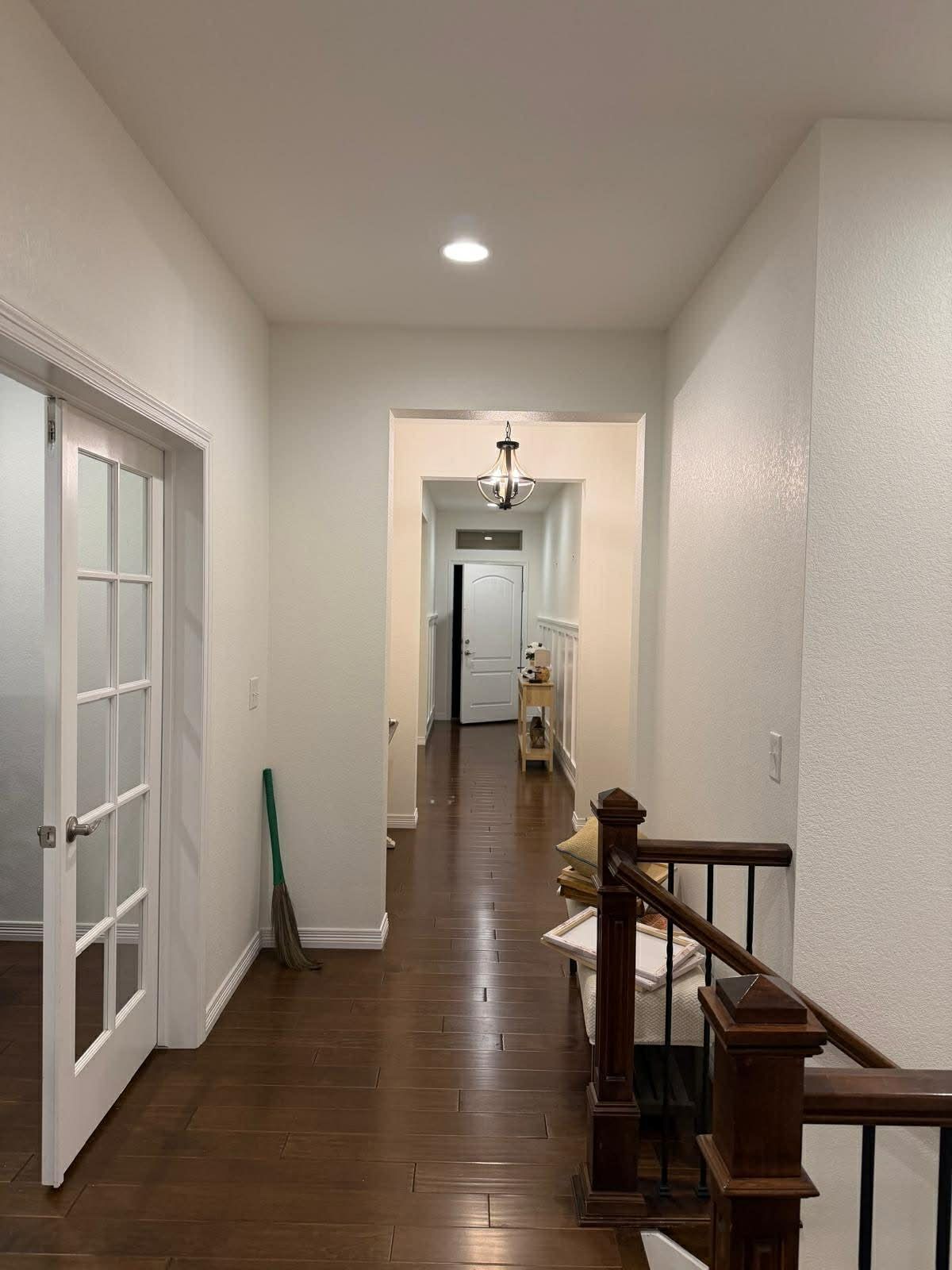 A brightly lit hallway with hardwood floors, white walls, and a wooden banister leading to an open door in the distance.