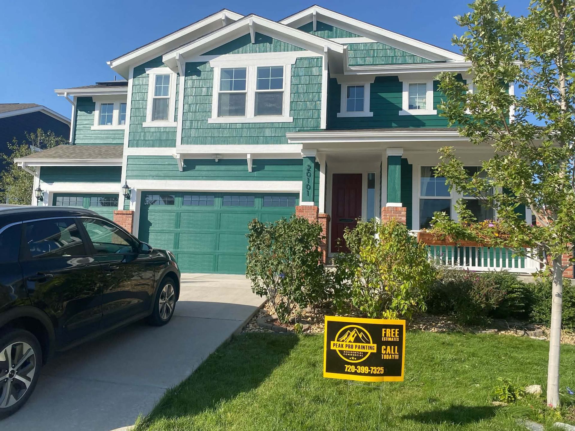 A teal, two-story house with a three-car garage and a small lawn featuring a yellow real estate sign in the front yard.