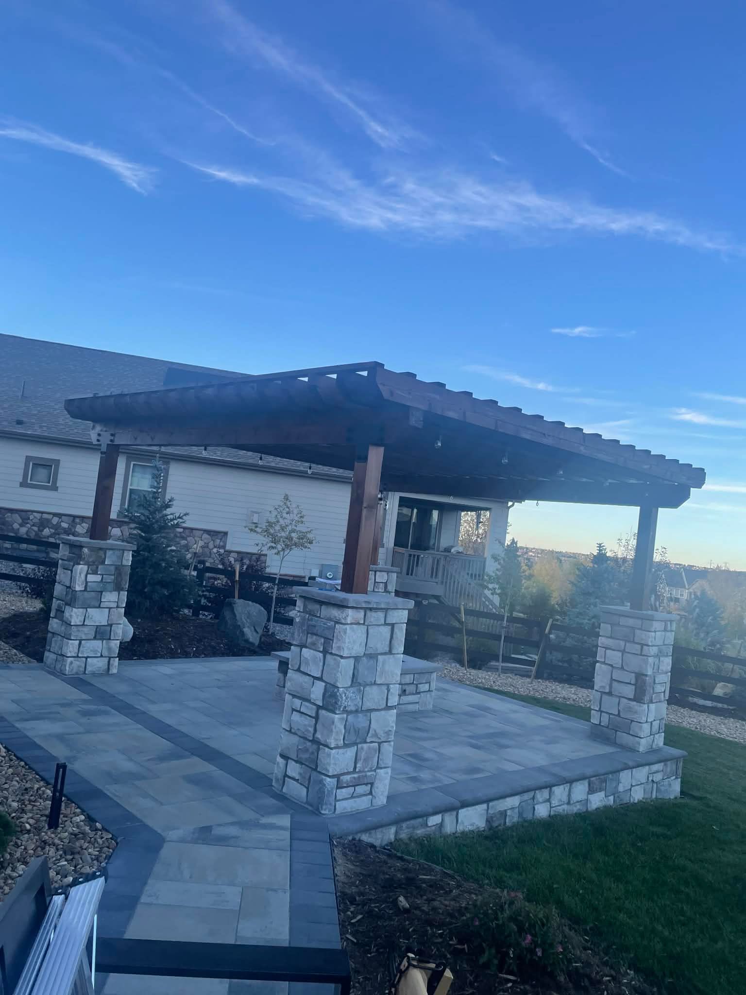 A backyard patio featuring a dark wood pergola supported by stone pillars on a paved stone surface under a blue sky.