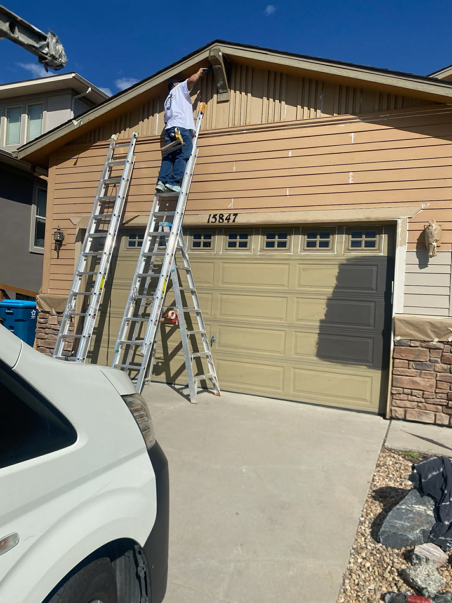 A person on a tall ladder uses a power tool to work on the exterior siding of a house above a garage.