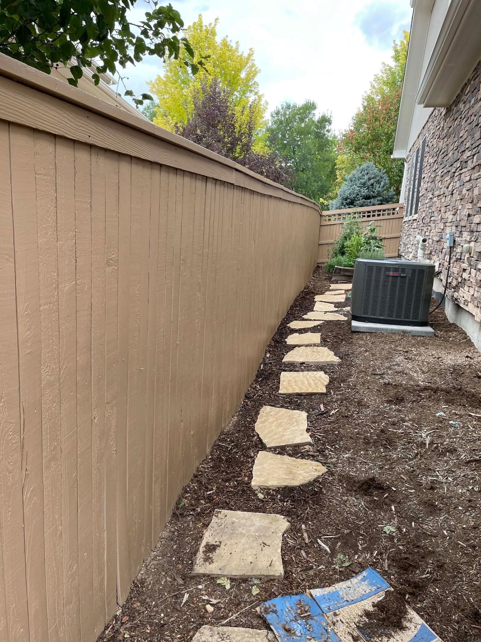 A stone stepping-stone path leads along a brown wooden privacy fence beside a brick house and an air conditioning unit.