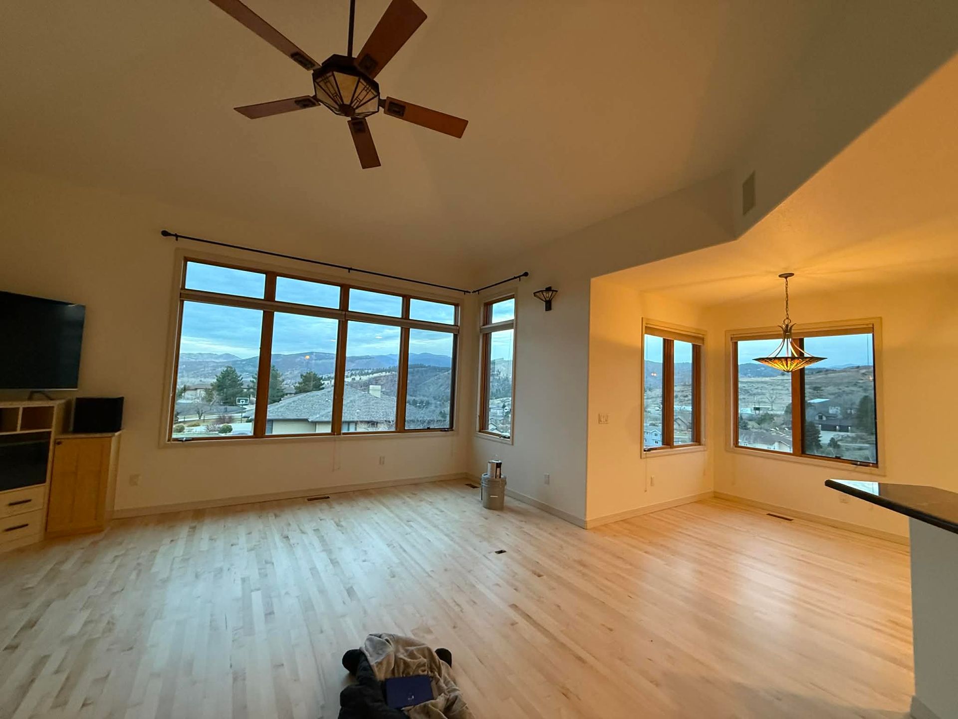 A wide-angle shot of a bright, open-concept living area with light wood floors, large windows, and a ceiling fan.