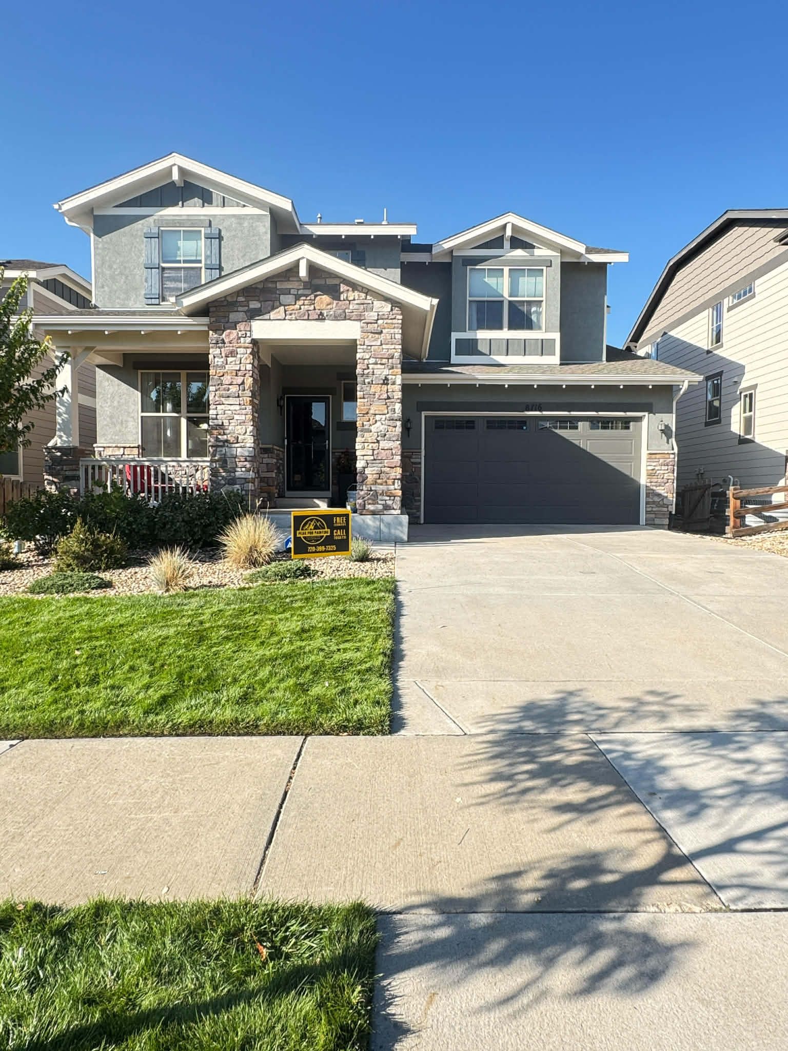 Two-story suburban house with gray siding, stone accents, a front porch, and a two-car garage under a clear blue sky.