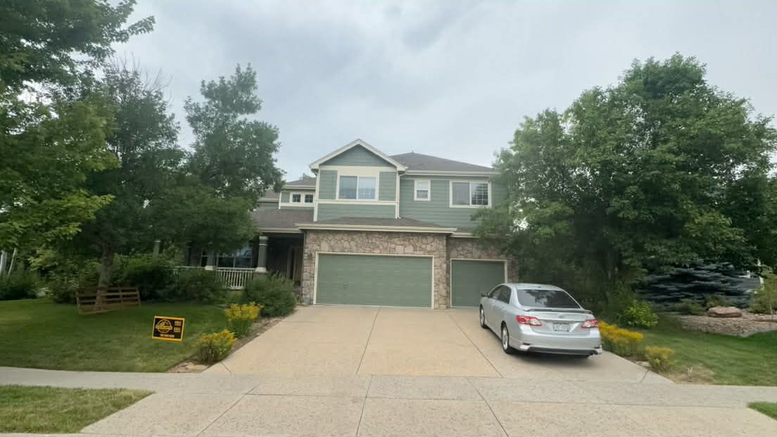 A two-story green suburban house with a stone-accented exterior, a three-car garage, and a silver car parked in the drive.