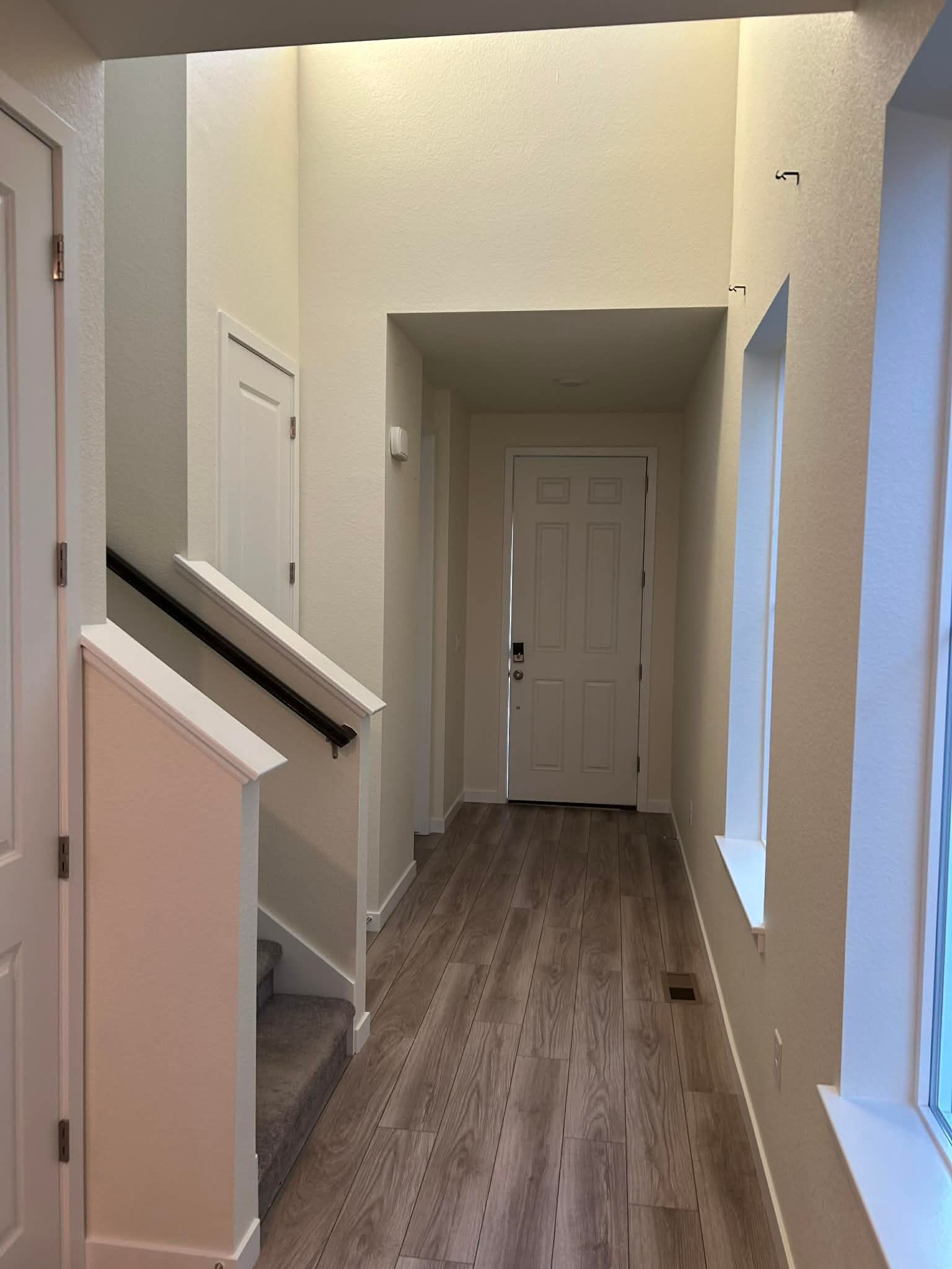 A hallway with wood-look flooring, light-colored walls, a staircase with a black handrail, and a white front door.